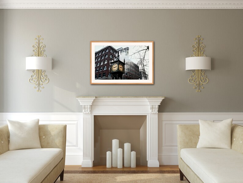 May include: A framed photograph of a clock tower in a city setting. The clock tower is brick with a gold clock face and is surrounded by other buildings. The photo is in a brown frame and is hanging on a gray wall.