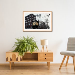 May include: A framed photograph of a brick building with a clock tower in the center. The clock face is visible and the building is surrounded by bare trees. The photo is hanging on a white wall above a wooden cabinet and a chair.