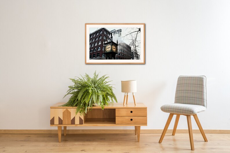 May include: A framed photograph of a brick building with a clock tower in the center. The clock face is visible and the building is surrounded by bare trees. The photo is hanging on a white wall above a wooden cabinet and a chair.