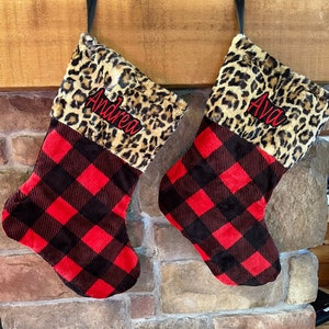May include: Two Christmas stockings with a red and black buffalo plaid pattern. The top portion of the stockings features a leopard print design. The name "Andrea" is embroidered on one stocking, and "Ava" is embroidered on the other.