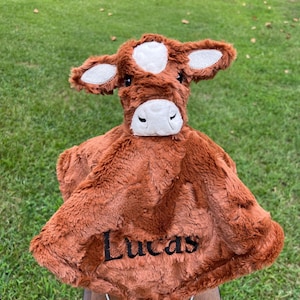 May include: A brown plush cow-shaped baby blanket with the name "Lucas" embroidered in black. The cow has white accents on the head and ears. The blanket is made of soft, fuzzy material.
