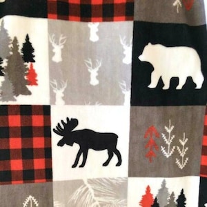 May include: A white fabric with a patchwork design featuring red and black plaid, moose, deer, bear, and pine tree silhouettes.
