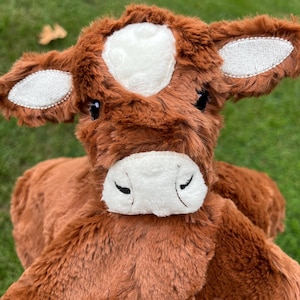 May include: A brown plush cow toy with a white patch on its head and white detailing on its ears. The soft toy has a white muzzle and closed black eyes. The stuffed animal is designed to resemble a farm animal, with a friendly expression.