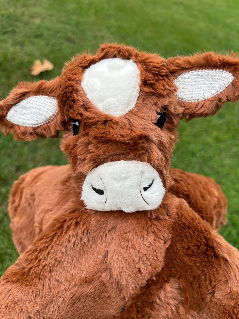 May include: A brown plush cow toy with a white patch on its head and white detailing on its ears. The soft toy has a white muzzle and closed black eyes. The stuffed animal is designed to resemble a farm animal, with a friendly expression.