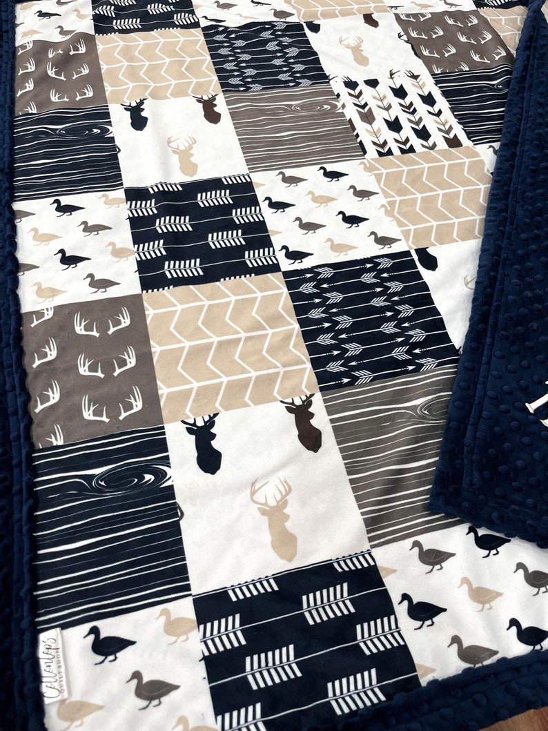 May include: A navy blue and tan baby blanket with a patchwork design featuring deer, arrows, and ducks. The blanket has a soft, plush texture.