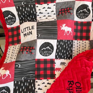 May include: A red and black patchwork quilt with a moose, bear, and forest-themed design. The quilt has the text "Little Man" and "Let's Sleep Under The Stars" on it. The quilt is paired with a red plush blanket with the text "Ollie Ryan" embroidered on it.