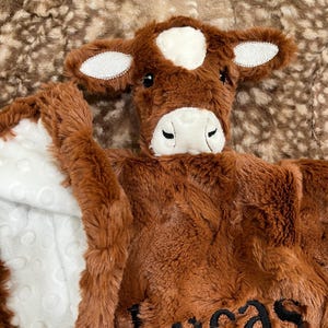 May include: A brown plush baby blanket with a cow face design. The blanket features a white patch on the forehead and ears, and the name "Lucas" embroidered in black. The blanket has a white, textured interior.