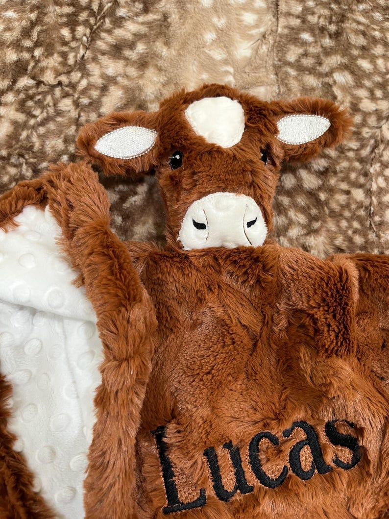 May include: A brown plush baby blanket with a cow face design. The blanket features a white patch on the forehead and ears, and the name "Lucas" embroidered in black. The blanket has a white, textured interior.