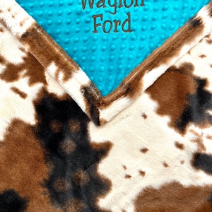 May include: A brown and white cow print baby blanket with a turquoise blue minky dot fabric on top. The minky fabric has the name "Waylon Ford" embroidered on it in brown thread.