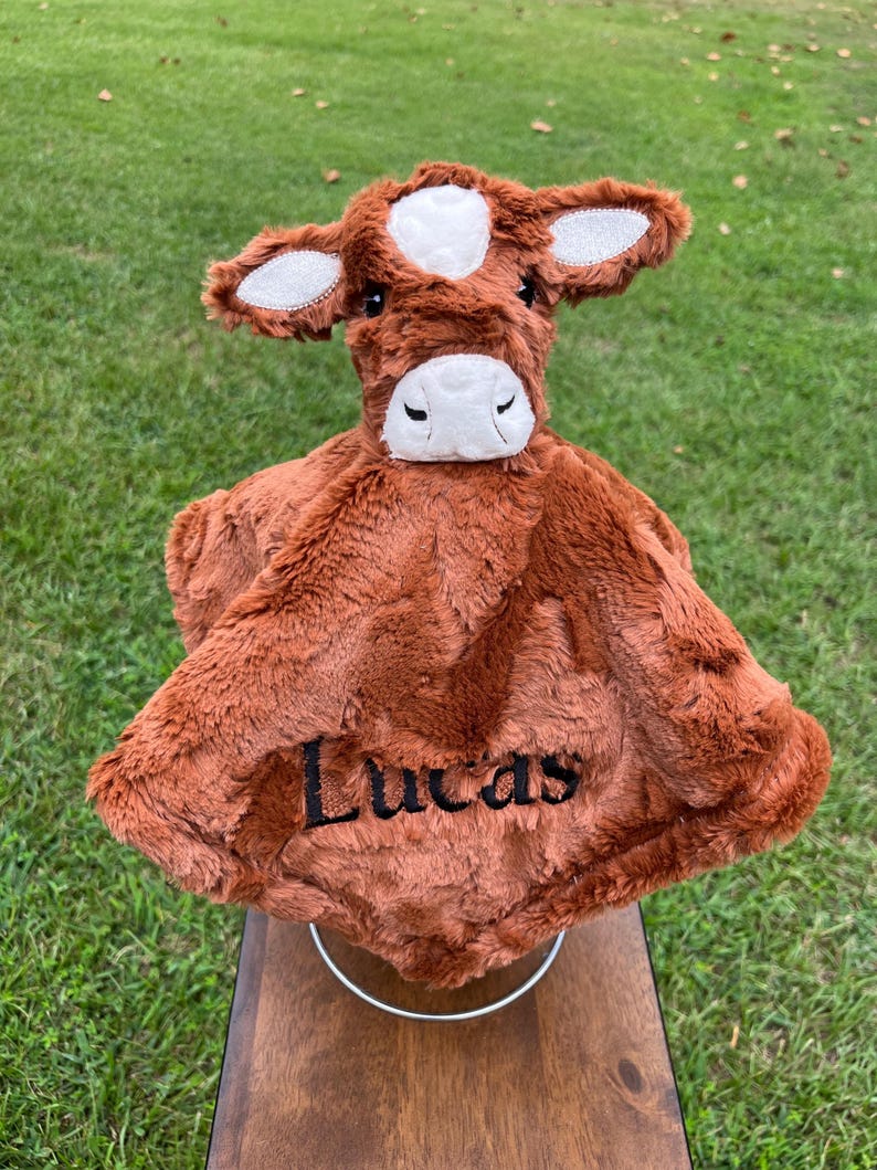May include: A brown plush cow-shaped baby blanket with the name "Lucas" embroidered in black. The cow has white accents on the head and ears. The blanket is made of soft, fuzzy material.