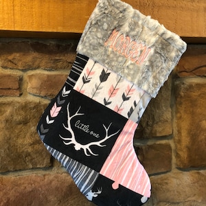 May include: A Christmas stocking with a faux fur cuff and a patchwork design. The stocking features a black and white deer antler graphic with the words "little one". The name "MORGEN" is embroidered in pink thread on the cuff.