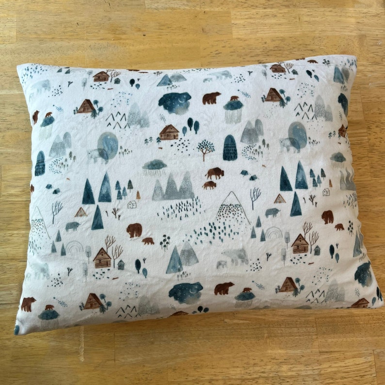 May include: A white pillowcase with a repeating pattern of brown bears, blue mountains, and small houses. The pattern is on a white background.