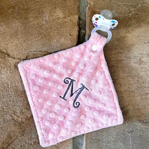 May include: Pink baby blanket with a pacifier clip and the letter M embroidered on it.