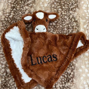 May include: A brown plush baby blanket with a cow head and the name "Lucas" embroidered on it. The blanket has a white, dotted lining and a soft, textured feel. The cow head features white ears and a white spot on its forehead.
