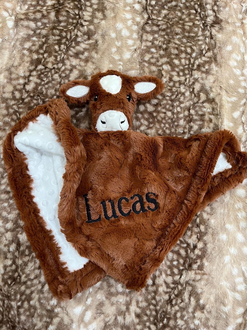 May include: A brown plush baby blanket with a cow head and the name "Lucas" embroidered on it. The blanket has a white, dotted lining and a soft, textured feel. The cow head features white ears and a white spot on its forehead.
