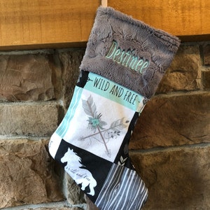 May include: A Christmas stocking with a gray plush cuff embroidered with the name "Destinee". The body of the stocking features a patchwork design with the words "WILD AND FREE", arrows, and a white horse silhouette.