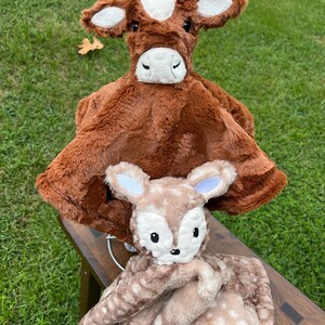 May include: Two plush baby security blankets. The top blanket is brown with a cow face, white spot, and sparkly ears. The bottom blanket is tan with a fawn face and a spotted pattern. Both blankets have soft, plush textures.