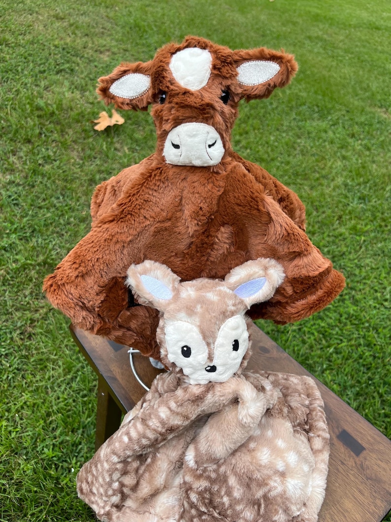 May include: Two plush baby security blankets. The top blanket is brown with a cow face, white spot, and sparkly ears. The bottom blanket is tan with a fawn face and a spotted pattern. Both blankets have soft, plush textures.