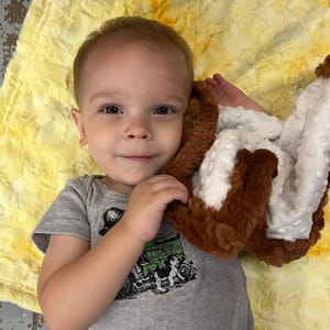 May include: A young child in a grey onesie with a green tractor design, lying on a yellow blanket. The child is holding a brown and white plush blanket, looking directly at the camera.