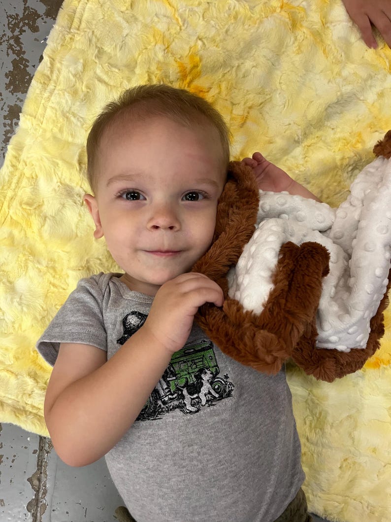May include: A young child in a grey onesie with a green tractor design, lying on a yellow blanket. The child is holding a brown and white plush blanket, looking directly at the camera.