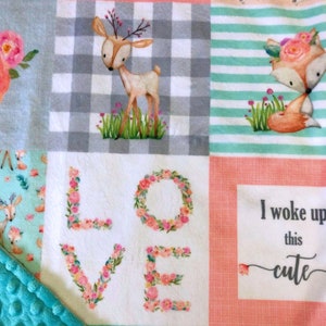 May include: A multi-colored baby blanket with a patchwork design. The blanket features a deer, a fox, and the words "LOVE" and "I woke up this cute".