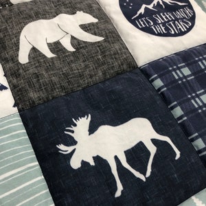May include: A patchwork quilt with a blue, gray, and white color scheme. The quilt features various animal silhouettes, including a bear, a moose, and a polar bear. The quilt also features a patch with the text "Let's Sleep Under The Stars".