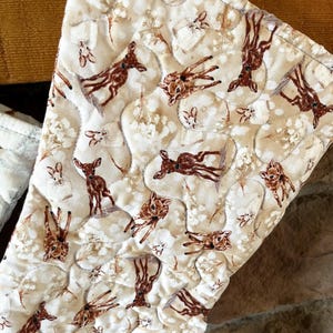 May include: Close-up of a quilted fabric with a repeating pattern of brown deer and white floral accents on a cream background. The fabric is stitched in a quilted pattern.