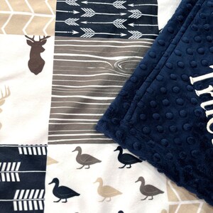May include: A multi-patterned baby blanket with a navy blue minky backing. The blanket features a variety of woodland animal and nature-inspired patterns, including deer, arrows, and ducks.