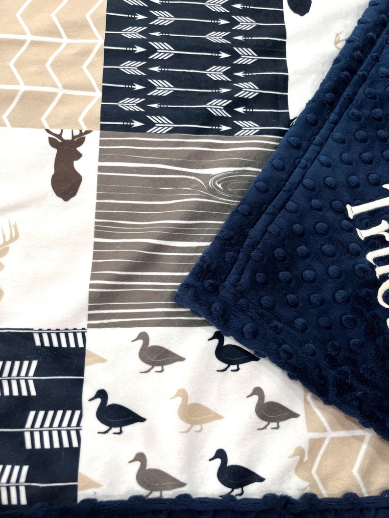 May include: A multi-patterned baby blanket with a navy blue minky backing. The blanket features a variety of woodland animal and nature-inspired patterns, including deer, arrows, and ducks.