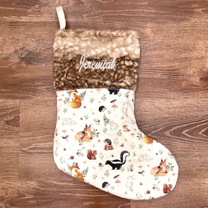 May include: A Christmas stocking with a cream-colored body featuring a woodland animal print. The top is brown faux fur, and the name "Jeremiah" is embroidered in white. A cream-colored ribbon loop is attached.