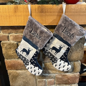 May include: Two Christmas stockings with gray faux fur tops and personalized names embroidered in black. The stockings feature a patchwork design with deer silhouettes and patterns in navy blue, white, and gray. Each stocking is hung by a white ribbon.