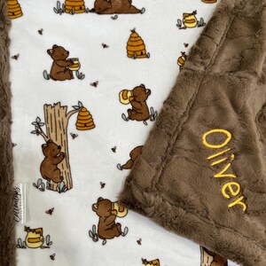 May include: A white baby blanket with a brown minky backing. The blanket features a repeating pattern of bears holding honey pots, beehives, and bees. The blanket is personalized with the name "Oliver" embroidered in yellow on the brown backing.