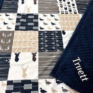 May include: A navy blue and tan patchwork baby blanket with a variety of woodland animal and arrow patterns. The blanket has a soft, plush backing with the name "Truett" embroidered on it.