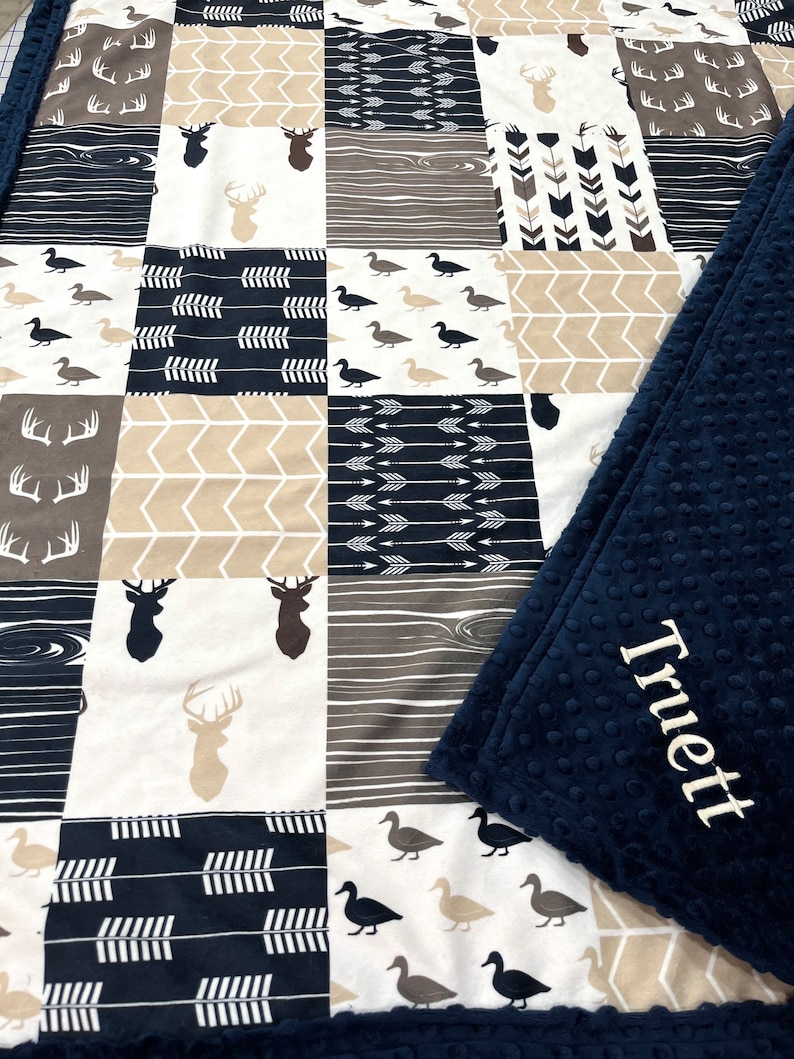 May include: A navy blue and tan patchwork baby blanket with a variety of woodland animal and arrow patterns. The blanket has a soft, plush backing with the name "Truett" embroidered on it.