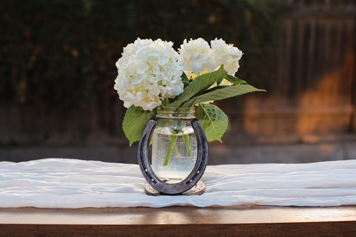 May include: A clear glass mason jar filled with water and white hydrangea flowers, held within a dark metal horseshoe frame that serves as a base. The arrangement features green leaves and is positioned upright on a flat surface.