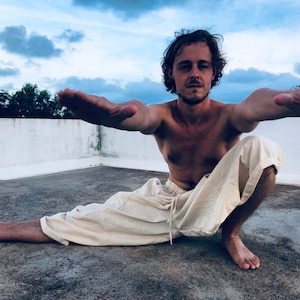 May include: A person is shown in a yoga pose, with arms outstretched and legs in a split. The person is wearing light-coloured trousers. The background is a cloudy sky.