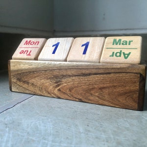 Peut inclure: Calendrier perpétuel en bois affichant "Mon 11 Mar". Le calendrier a une base en bois brun foncé et des blocs de bois individuels pour le jour, le mois et la date. Les chiffres et le texte sont en rouge, bleu et vert.