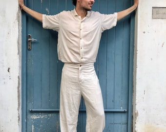 Puede incluir: Una persona vestida con una camisa de lino color crema y pantalones a juego. La camisa tiene mangas cortas y botones. Los pantalones son de pierna ancha. La persona lleva sandalias marrones. El fondo es una puerta azul.