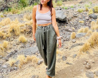May include: A light grey tank top and olive green linen pants with a tie waist. The pants have a wide leg design. The person is standing on a rocky path with dry grass and rocks.