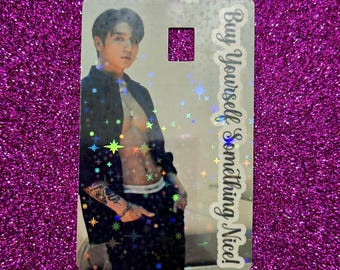 May include: A holographic card featuring a person in a black jacket and pants. The card has a rectangular cutout and text that reads "Buy Yourself Something Nice!" The card is decorated with star-shaped holographic accents.