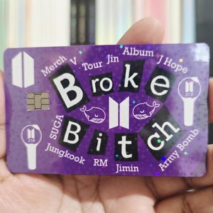 May include: A purple card with the words "Broke Bitch" in a bold font. The card has various words and symbols, including "Merch," "Album," and "Army Bomb." The card is held in a hand.