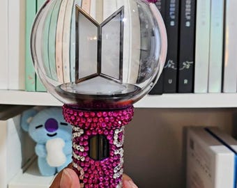 Può includere: Un bastoncino luminoso sferico in plastica trasparente con un logo BTS argentato al centro, sormontato da una piccola statuetta dai capelli castani. L'impugnatura e la base della sfera sono ricoperte da densi e scintillanti strass rosa e bianchi.