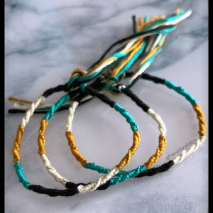 May include: Three handmade bracelets with a braided design. The bracelets feature sections of black, teal, gold, and white thread. The bracelets are tied together with matching threads. The bracelets are a type of jewelry.
