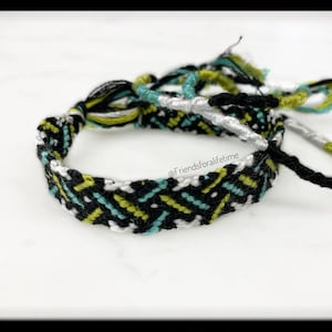 May include: A black, white, green, and blue friendship bracelet with a chevron pattern. The bracelet is tied in a knot and has frayed ends.
