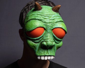 May include: A green monster mask with orange eyes and horns. The mask has a textured surface and a toothy grin.