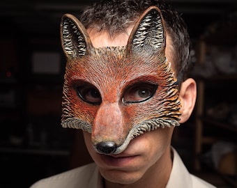 May include: A close-up of a person wearing a realistic fox mask. The mask is made of brown, orange, and black faux fur and has a detailed snout and ears.