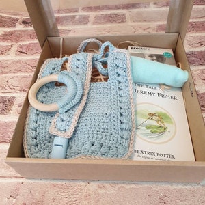 May include: A blue and beige crocheted baby bib with a wooden teething ring, a blue teething toy, and a copy of "The Tale of Jeremy Fisher" by Beatrix Potter in a brown gift box.