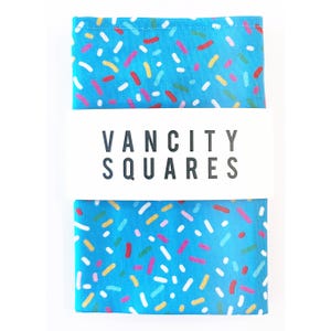 Könnte beinhalten: Ein blauer Stoffquadrat mit weißem Hintergrund und einem Muster aus bunten Streuseln. Der Stoffquadrat hat den Text "VANCITY SQUARES" aufgedruckt.