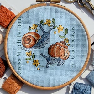 Peut inclure: Motif de point de croix avec deux escargots aux coquilles marron et bleues, entourés de fleurs jaunes et de feuilles vertes, sur fond bleu clair. Le texte indique "Cross Stitch Pattern" et "Fifi Grace Designs".