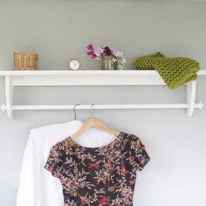 May include: White wooden shelf with a white metal rod for hanging clothes. A green knitted scarf is on the shelf. A white shirt and a brown and black floral dress are hanging on the rod.